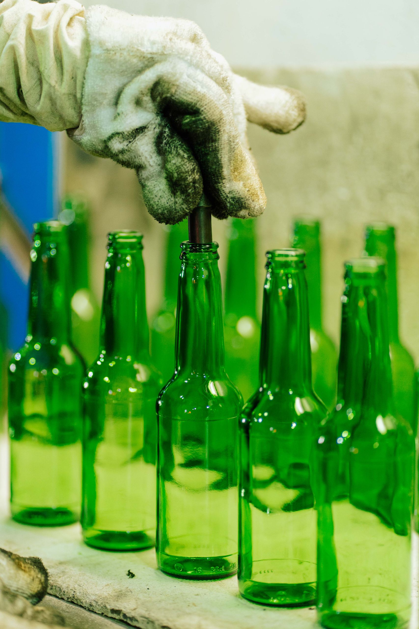 A gloved worker handles green glass bottles in a factory setting.